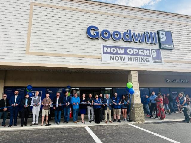 Group of people cutting a blue ribbon outside a Goodwill store during a grand opening event with balloons nearby.