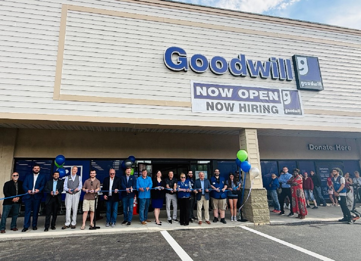 Group of people cutting a blue ribbon outside a Goodwill store during a grand opening event with balloons nearby.