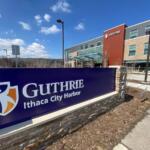 Front sign for Guthrie Ithaca City Harbor with a modern medical campus building in the background under a blue sky.