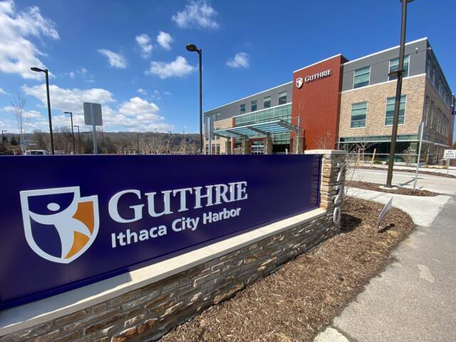 Front sign for Guthrie Ithaca City Harbor with a modern medical campus building in the background under a blue sky.