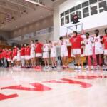 Basketball team in red and white uniforms stands in a line on a gym court before a game or ceremony, with red bleachers in the background.