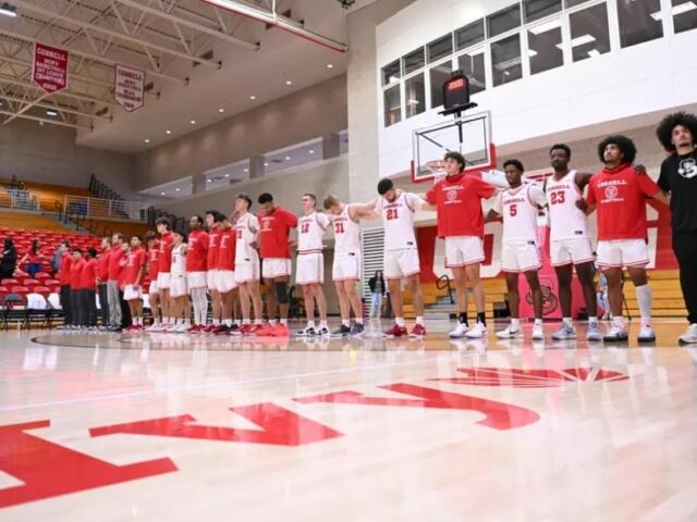 Basketball team in red and white uniforms stands in a line on a gym court before a game or ceremony, with red bleachers in the background.