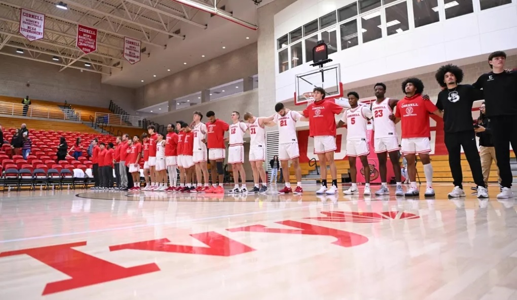 Basketball team in red and white uniforms stands in a line on a gym court before a game or ceremony, with red bleachers in the background.