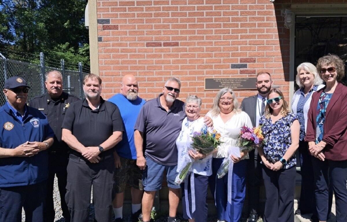 Group of ten adults posing outside a brick building; two women in the center hold bouquets and a card/paper.