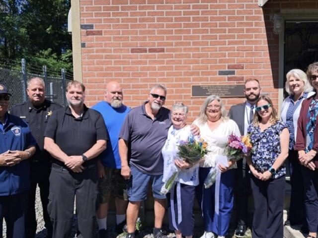 Group of ten adults posing outside a brick building; two women in the center hold bouquets and a card/paper.