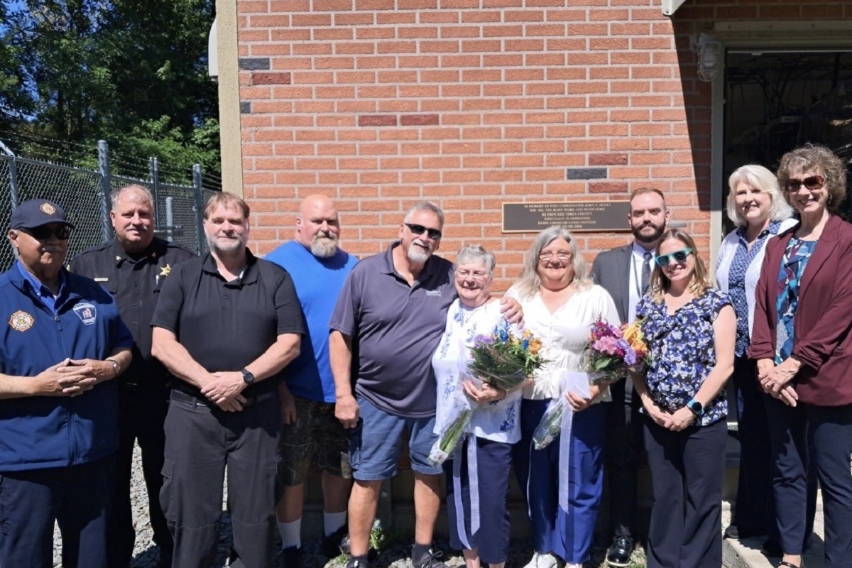 Group of ten adults posing outside a brick building; two women in the center hold bouquets and a card/paper.