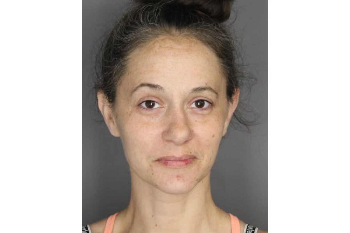 Front-facing head-and-shoulders portrait of a woman with light skin, dark hair in a messy bun, neutral expression and visible facial redness/acne.