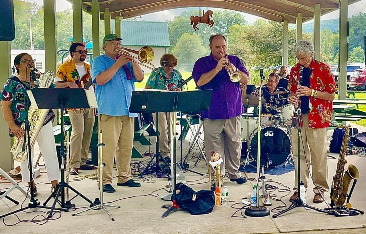 Outdoor brass band performs under a pavilion, players in colorful shirts with trumpets, trombone, saxophone, and drums nearby.