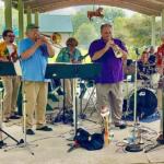 Outdoor brass band performs under a pavilion, players in colorful shirts with trumpets, trombone, saxophone, and drums nearby.