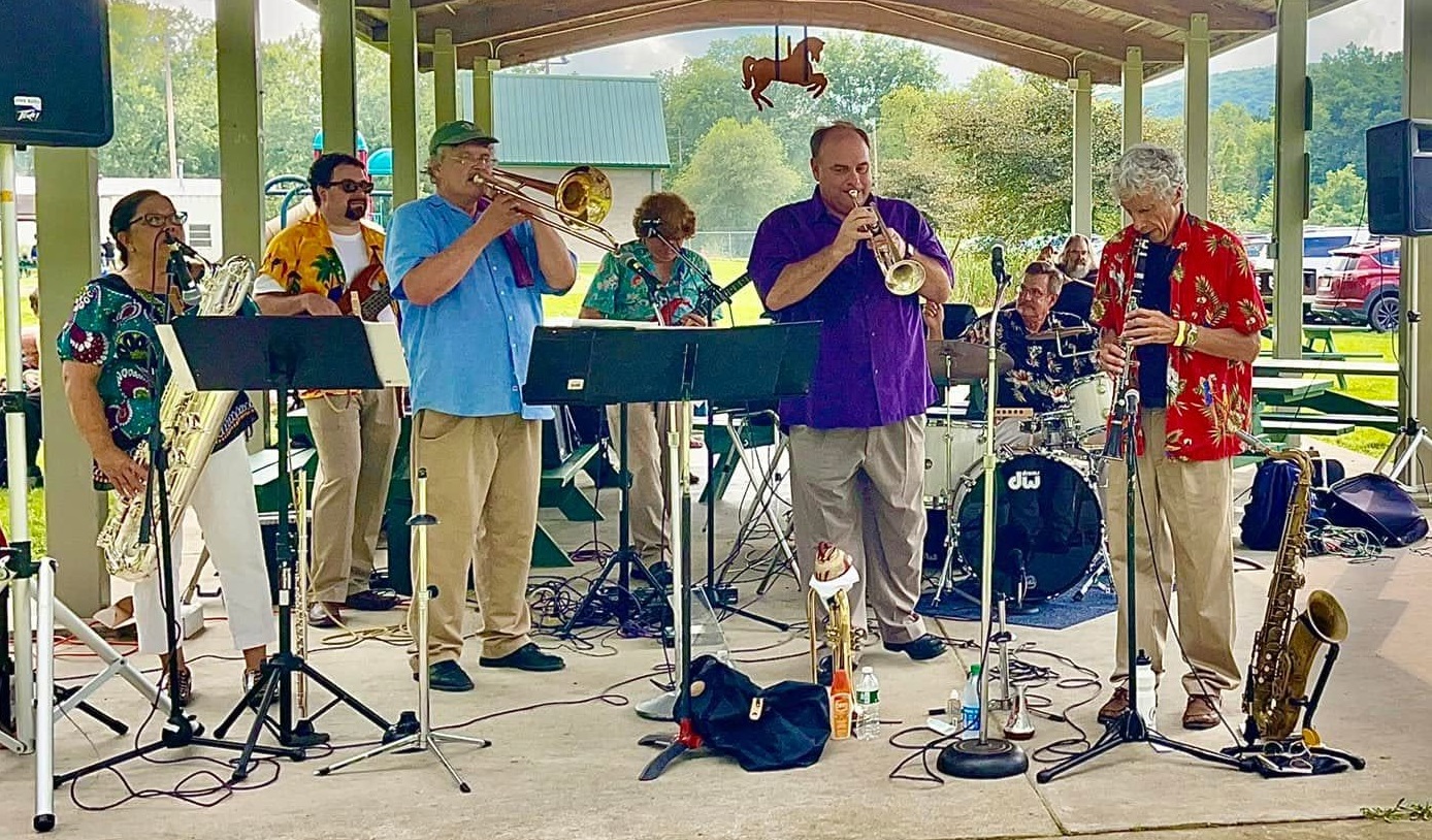 Outdoor brass band performs under a pavilion, players in colorful shirts with trumpets, trombone, saxophone, and drums nearby.