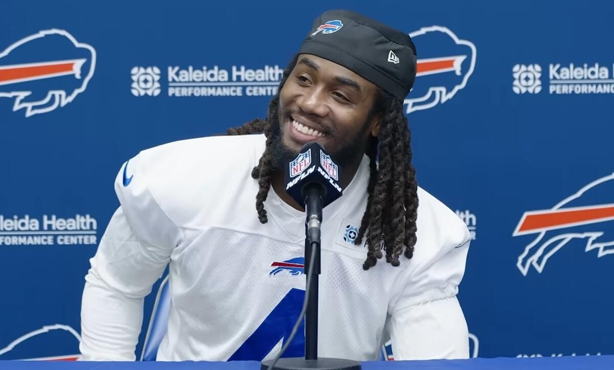 Bills player at a press conference, smiling into a microphone with a blue backdrop featuring Kaleida Health logos.