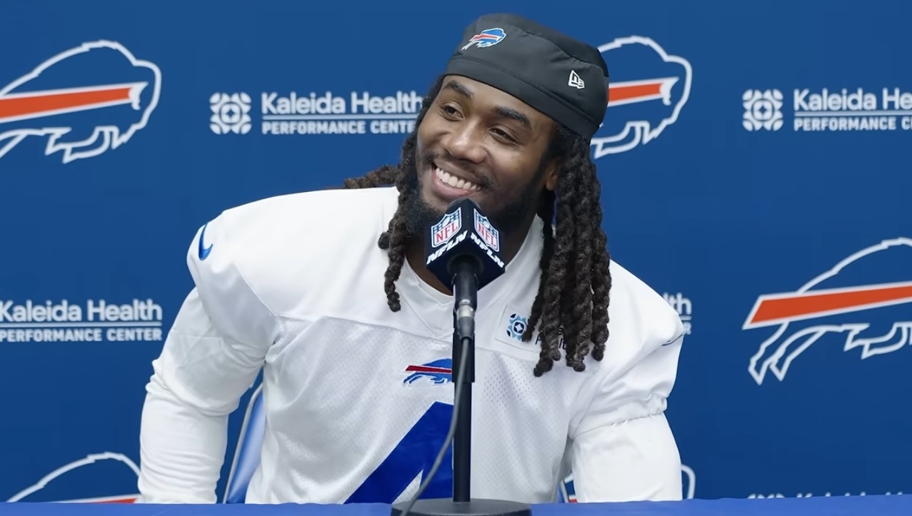 Bills player at a press conference, smiling into a microphone with a blue backdrop featuring Kaleida Health logos.