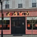 Pink storefront for 'maru korea' with black doors, large front windows showing orange chairs inside and a neon 'open' sign above.