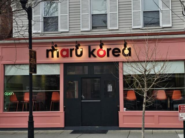 Pink storefront for 'maru korea' with black doors, large front windows showing orange chairs inside and a neon 'open' sign above.