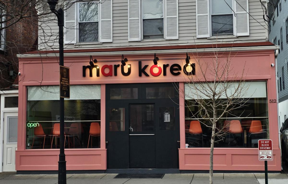 Pink storefront for 'maru korea' with black doors, large front windows showing orange chairs inside and a neon 'open' sign above.