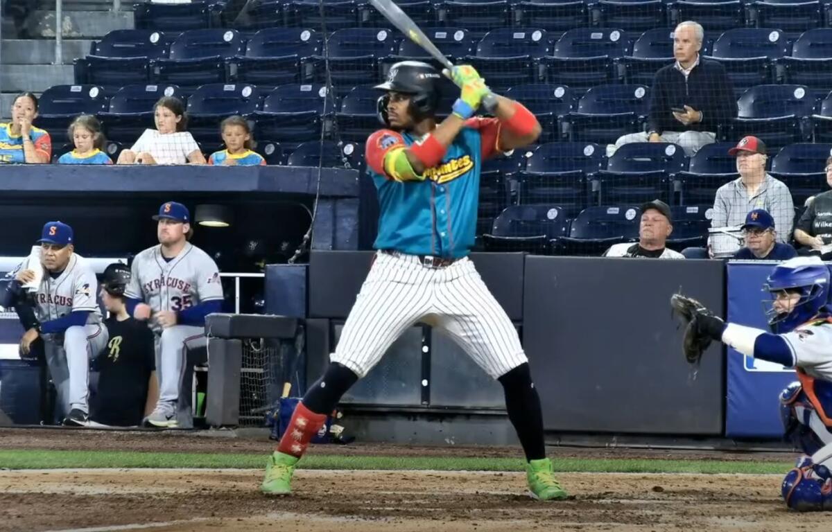 Baseball batter in teal jersey swings the bat at home plate, catcher ready behind him and fans watching in the stands behind.