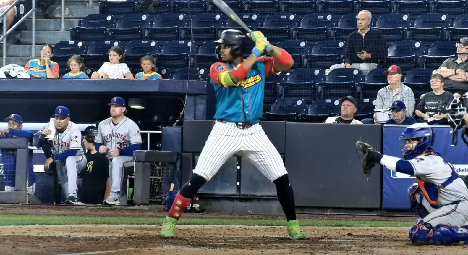 Baseball batter in teal jersey swings the bat at home plate, catcher ready behind him and fans watching in the stands behind.