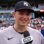 Baseball player in Yankees cap being interviewed on the field with a YES microphone; stadium crowd in the background.