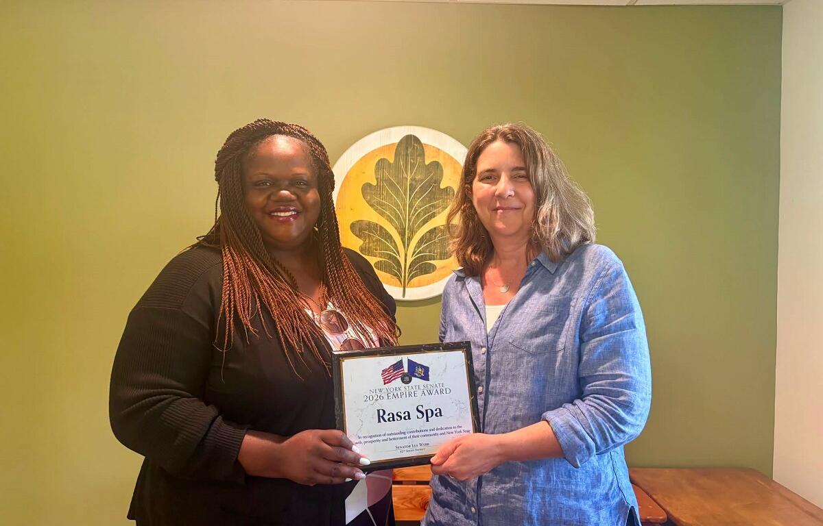 Two smiling women stand indoors as one presents a framed certificate reading 'Rasa Spa' during an award presentation in a green-walled room with a decorative emblem.