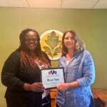 Two smiling women stand indoors as one presents a framed certificate reading 'Rasa Spa' during an award presentation in a green-walled room with a decorative emblem.