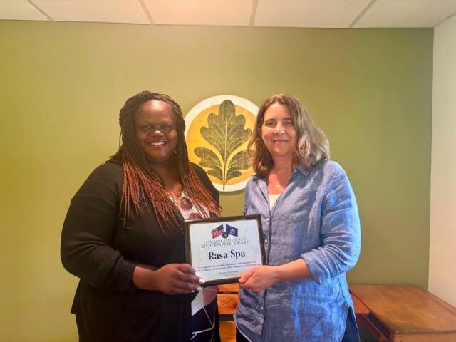 Two smiling women stand indoors as one presents a framed certificate reading 'Rasa Spa' during an award presentation in a green-walled room with a decorative emblem.