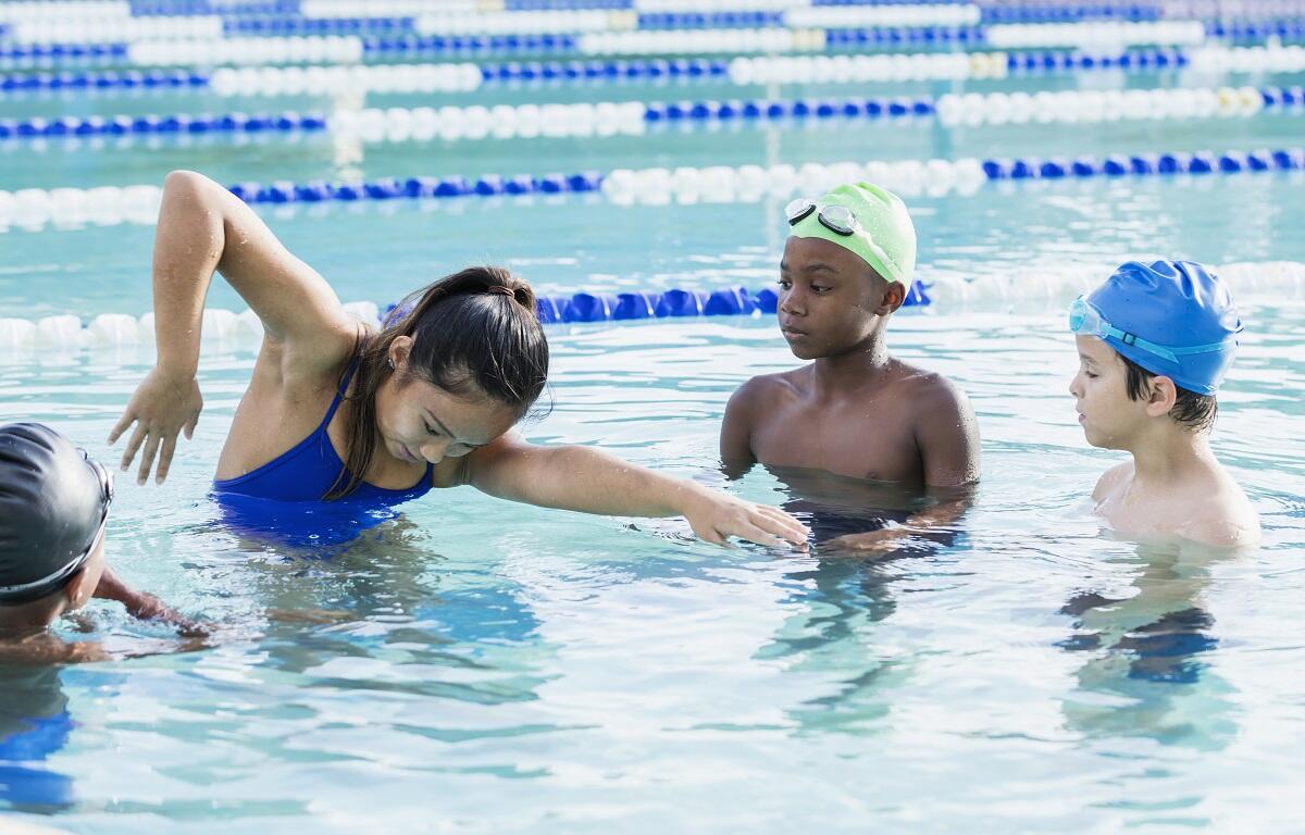 Group of children in a swimming lesson, a girl in a blue swimsuit reaching forward in the water while two boys watch nearby.