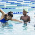 Group of children in a swimming lesson, a girl in a blue swimsuit reaching forward in the water while two boys watch nearby.
