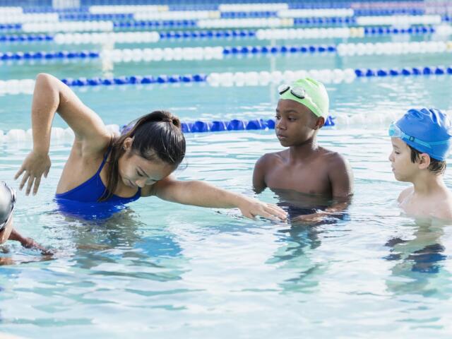 Group of children in a swimming lesson, a girl in a blue swimsuit reaching forward in the water while two boys watch nearby.