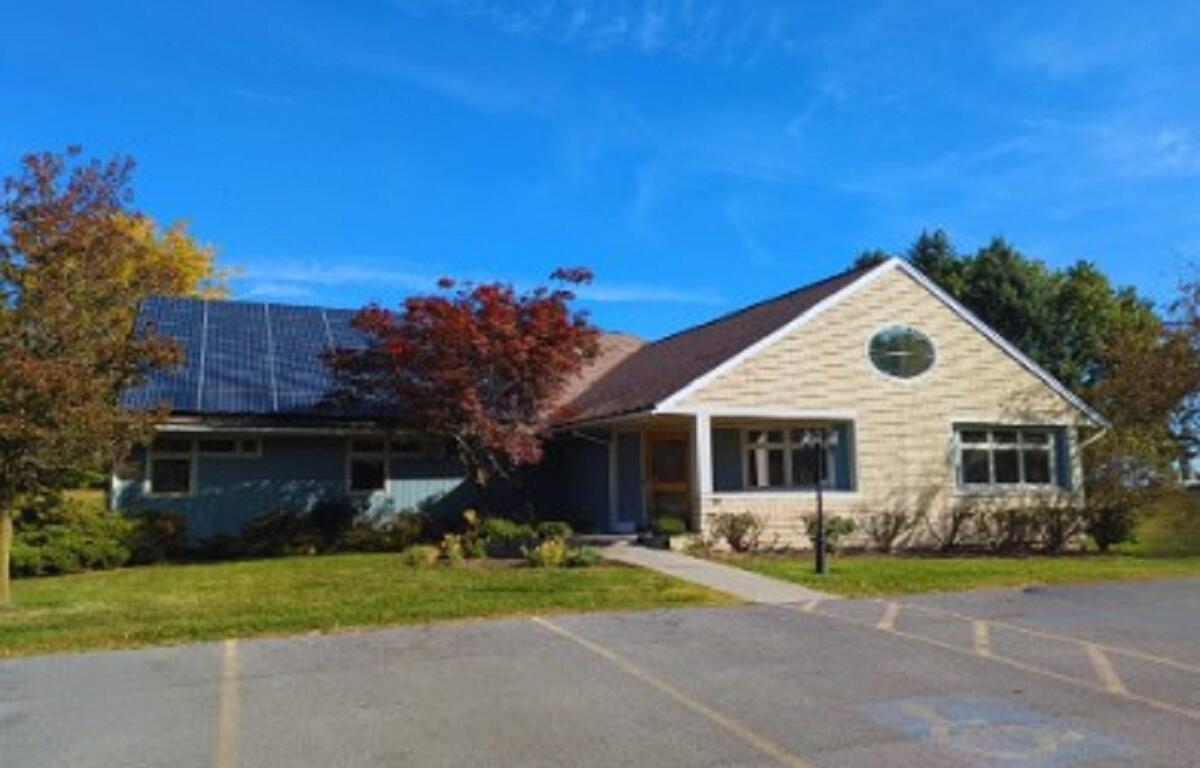 Front view of a single-story building with solar panels on the left roof, blue sky, and surrounding trees in autumn colors.