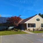 Front view of a single-story building with solar panels on the left roof, blue sky, and surrounding trees in autumn colors.