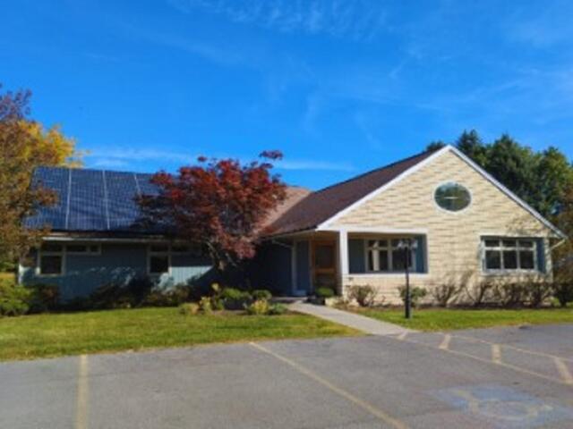Front view of a single-story building with solar panels on the left roof, blue sky, and surrounding trees in autumn colors.