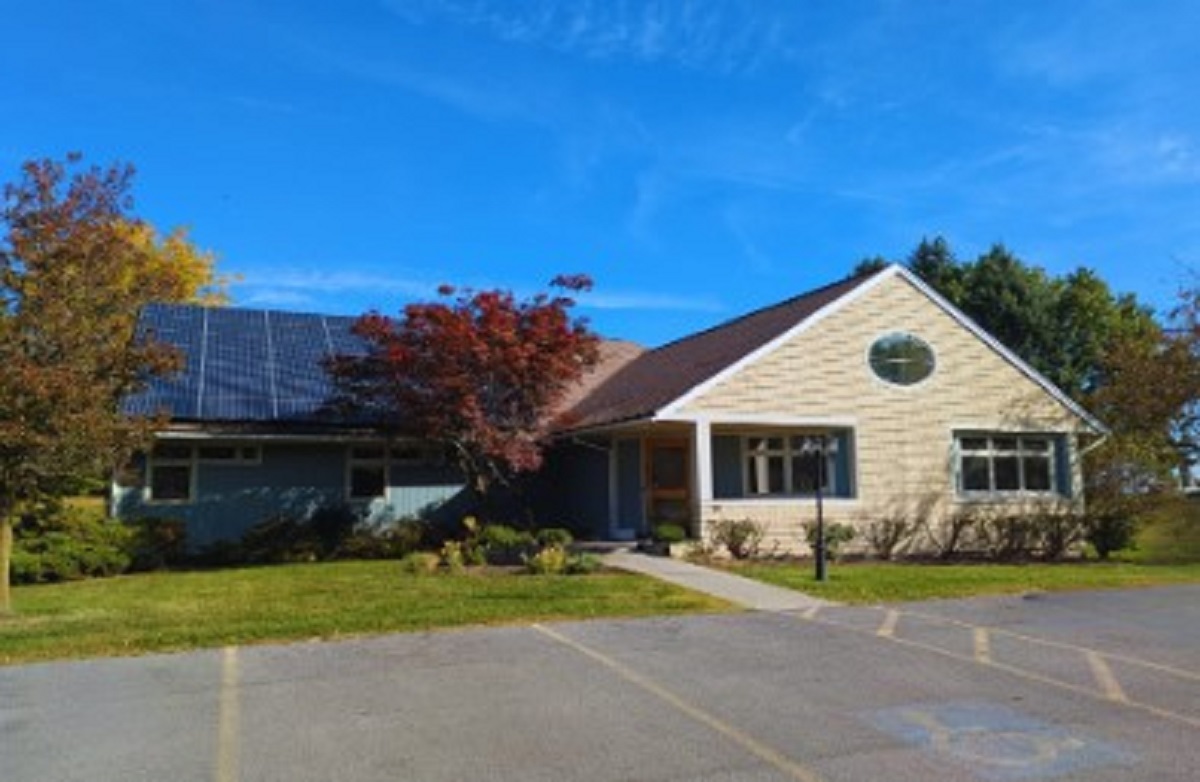 Front view of a single-story building with solar panels on the left roof, blue sky, and surrounding trees in autumn colors.