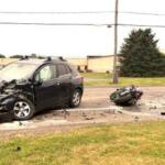 Black SUV with severe front-end damage after a collision on a road, debris scattered across the street, and a toppled motorcycle nearby.