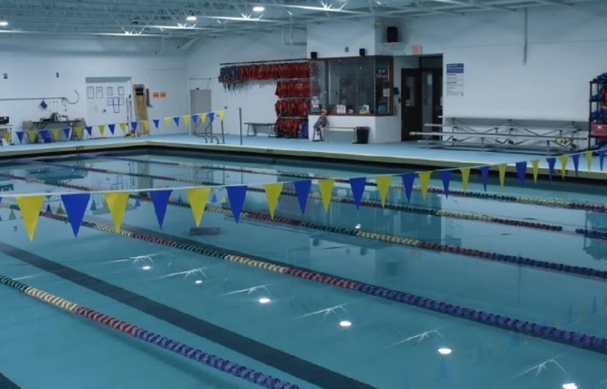 Indoor swimming pool with lane markers and yellow/blue pennant flags overhead, lifeguard station in background.