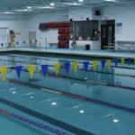 Indoor swimming pool with lane markers and yellow/blue pennant flags overhead, lifeguard station in background.
