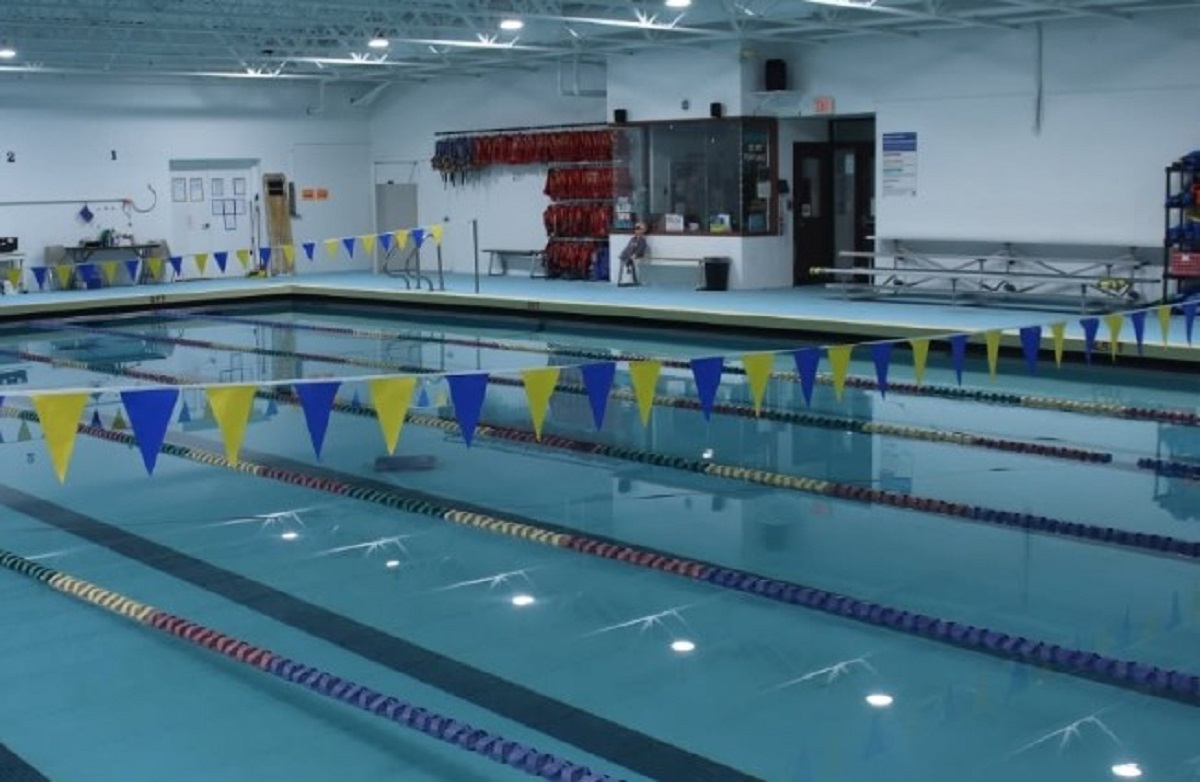 Indoor swimming pool with lane markers and yellow/blue pennant flags overhead, lifeguard station in background.